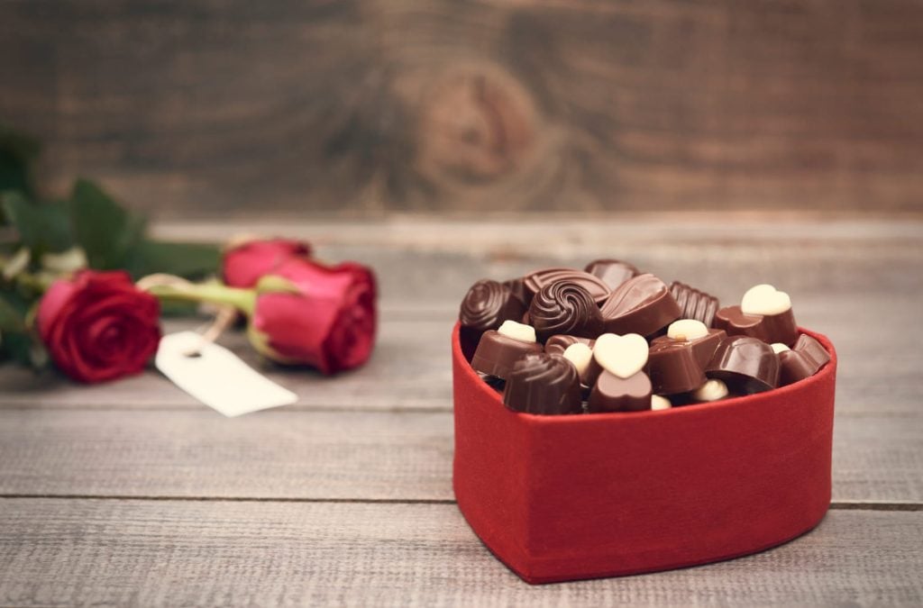 Caixa vermelha com chocolates em formato de coração e rosas vermelhas ao fundo.