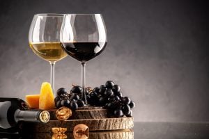 Taças de vinho tinto e branco ao lado de uvas pretas, queijo, nozes e garrafa sobre tábua de madeira.