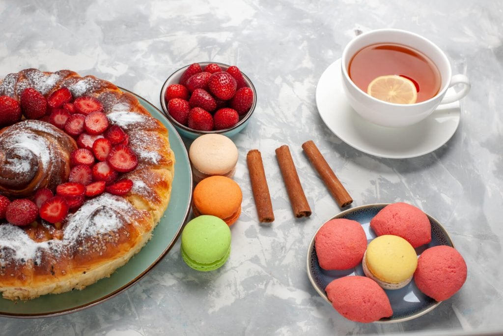 Mesa posta com rosca doce coberta com morangos e açúcar de confeiteiro, xícara de chá com limão, macarons e doces sobre superfície clara.