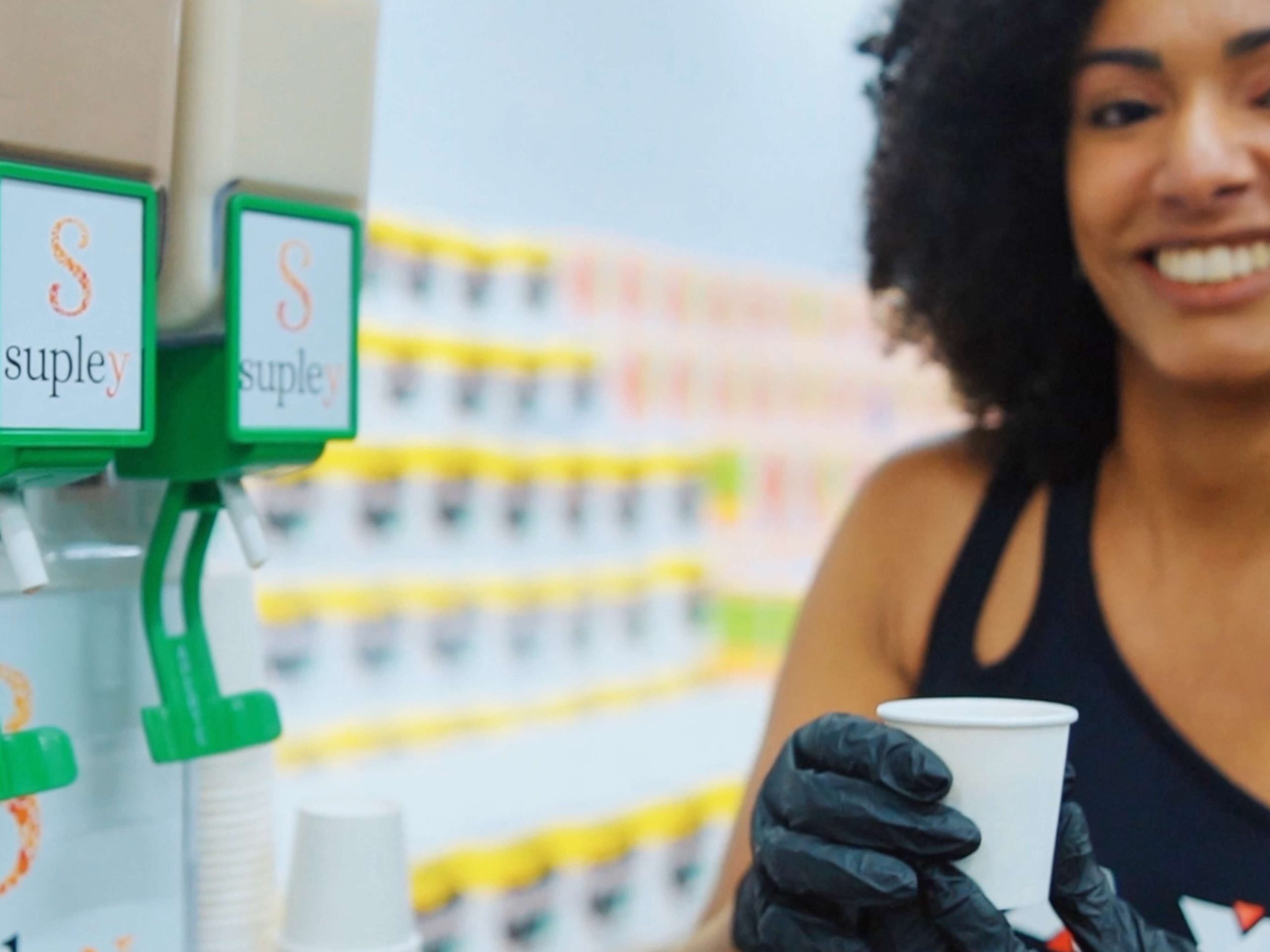 Mulher sorrindo serve bebida em copinho durante degustação da Supley em loja do Sam’s Club