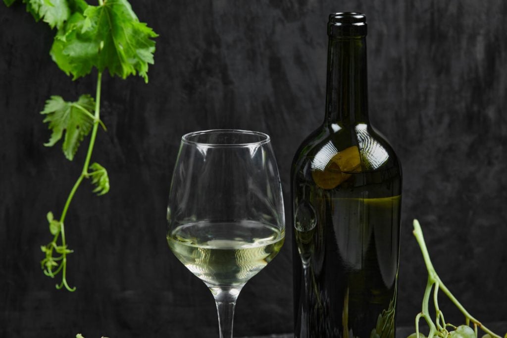 Taça e garrafa de vinho branco sobre superfície escura, com folhas de videira e uvas verdes ao redor.