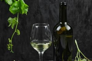 Taça e garrafa de vinho branco sobre superfície escura, com folhas de videira e uvas verdes ao redor.