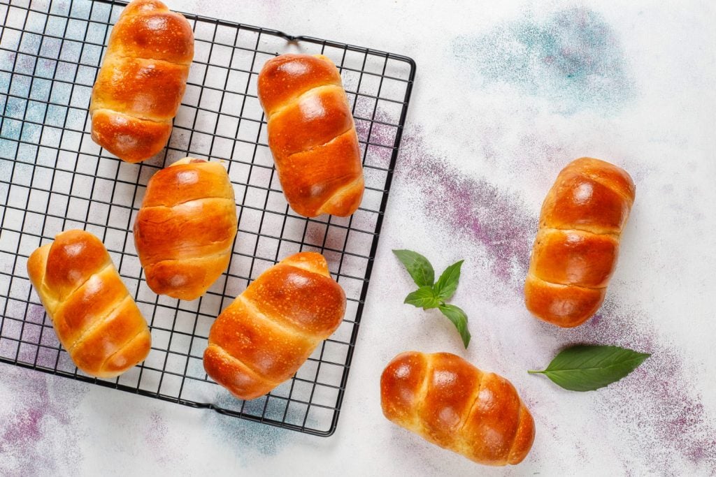 Pães brioche dourados sobre grade de resfriamento em bancada clara, com folhas verdes decorativas ao lado.