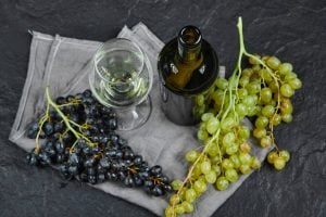 Garrafa e taça de vinho branco ao lado de cachos de uvas verdes e roxas sobre pano cinza e superfície escura.