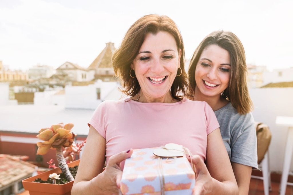 Mulher sorrindo recebe presente embrulhado em terraço ao ar livre, enquanto uma jovem a abraça por trás em momento de celebração.