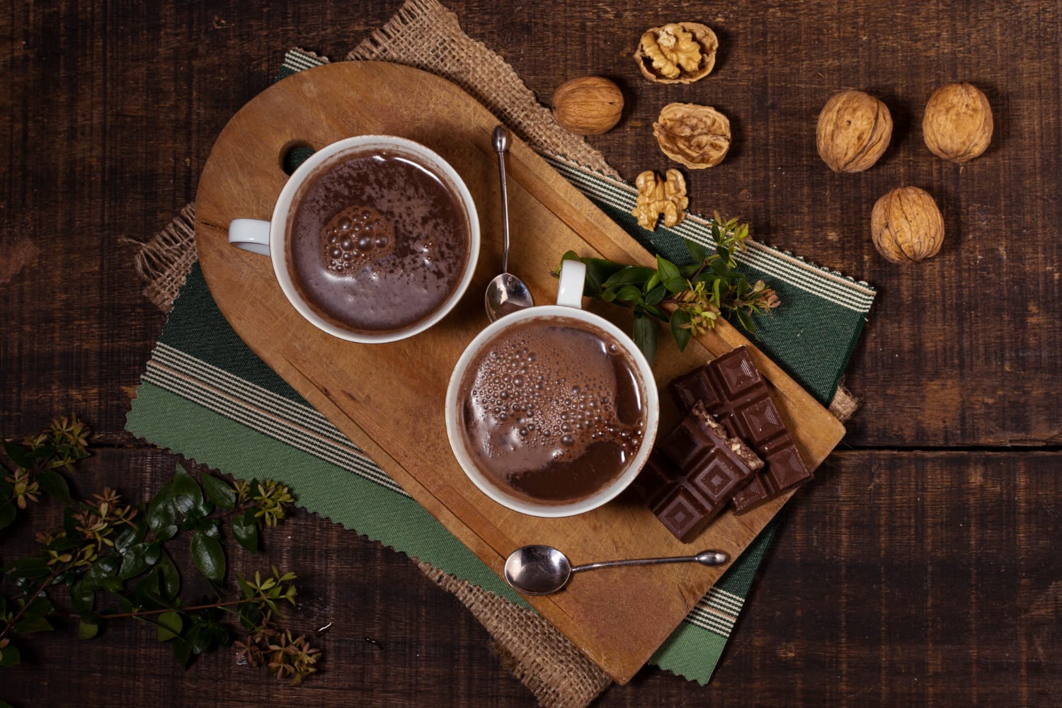 Duas xícaras de chocolate quente servidas com barra de chocolate e nozes sobre tábua de madeira.